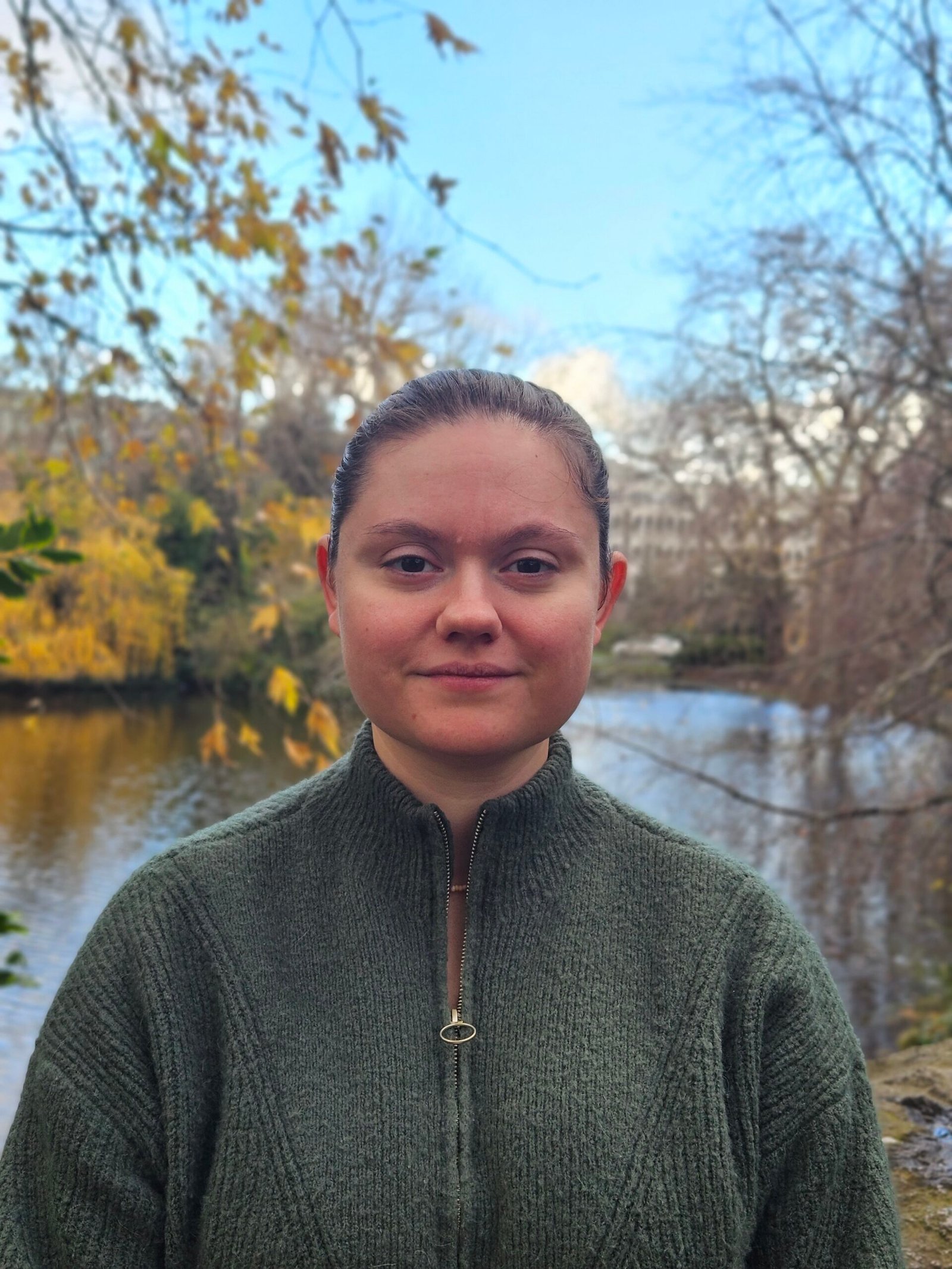 Portrait professionnel d'Angeline Staiquly souriante, vêtue d'un pull vert, avec un paysage naturel en arrière-plan.